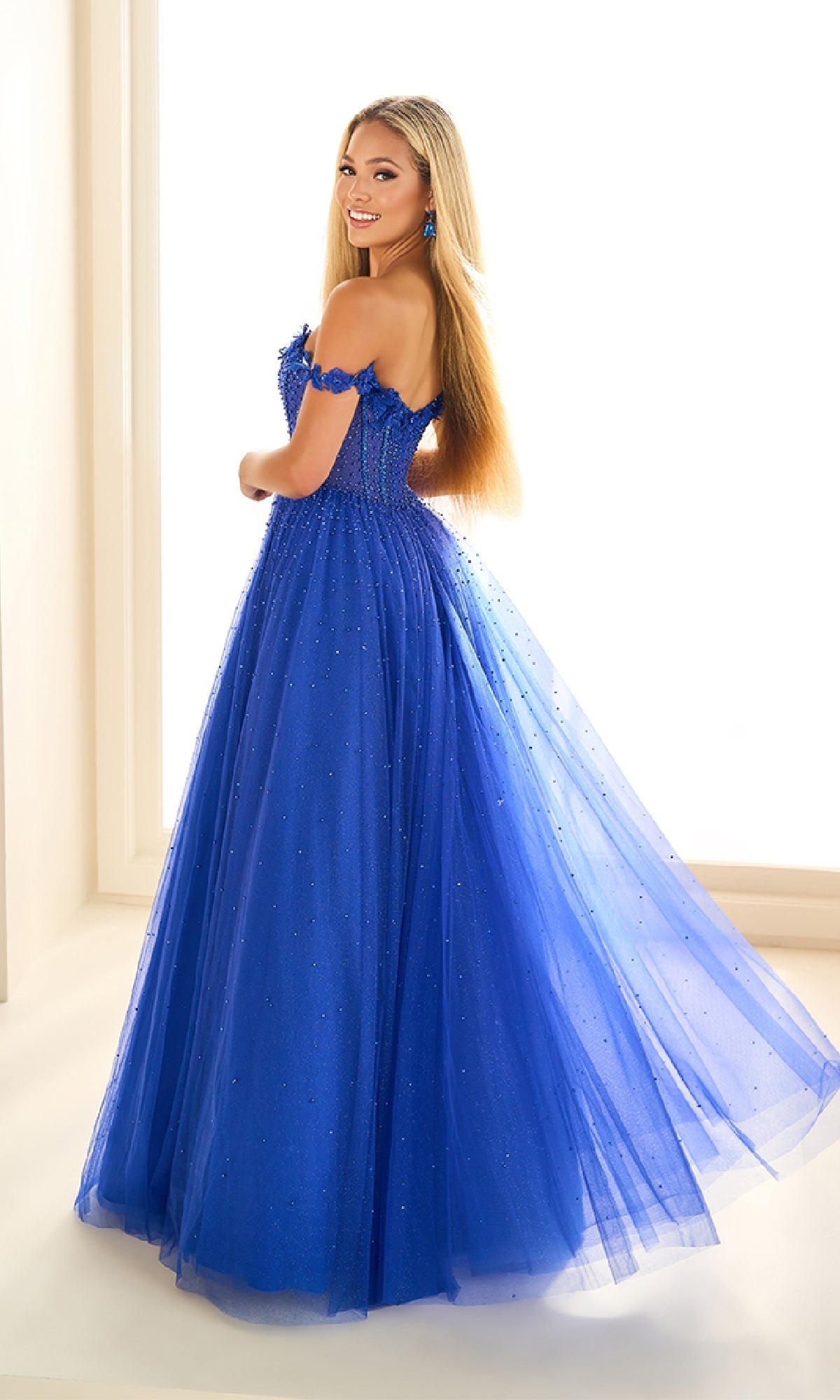 A woman stands in a well-lit room wearing the Ellie Wilde EW36071 long prom dress, an off-the-shoulder ball gown with beaded embellishments, looking back over her shoulder and smiling.